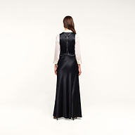A woman with long brown hair stands facing away, wearing the Vespera Suspender Maxi Skirt in black satin over a white blouse. The background is plain and white.