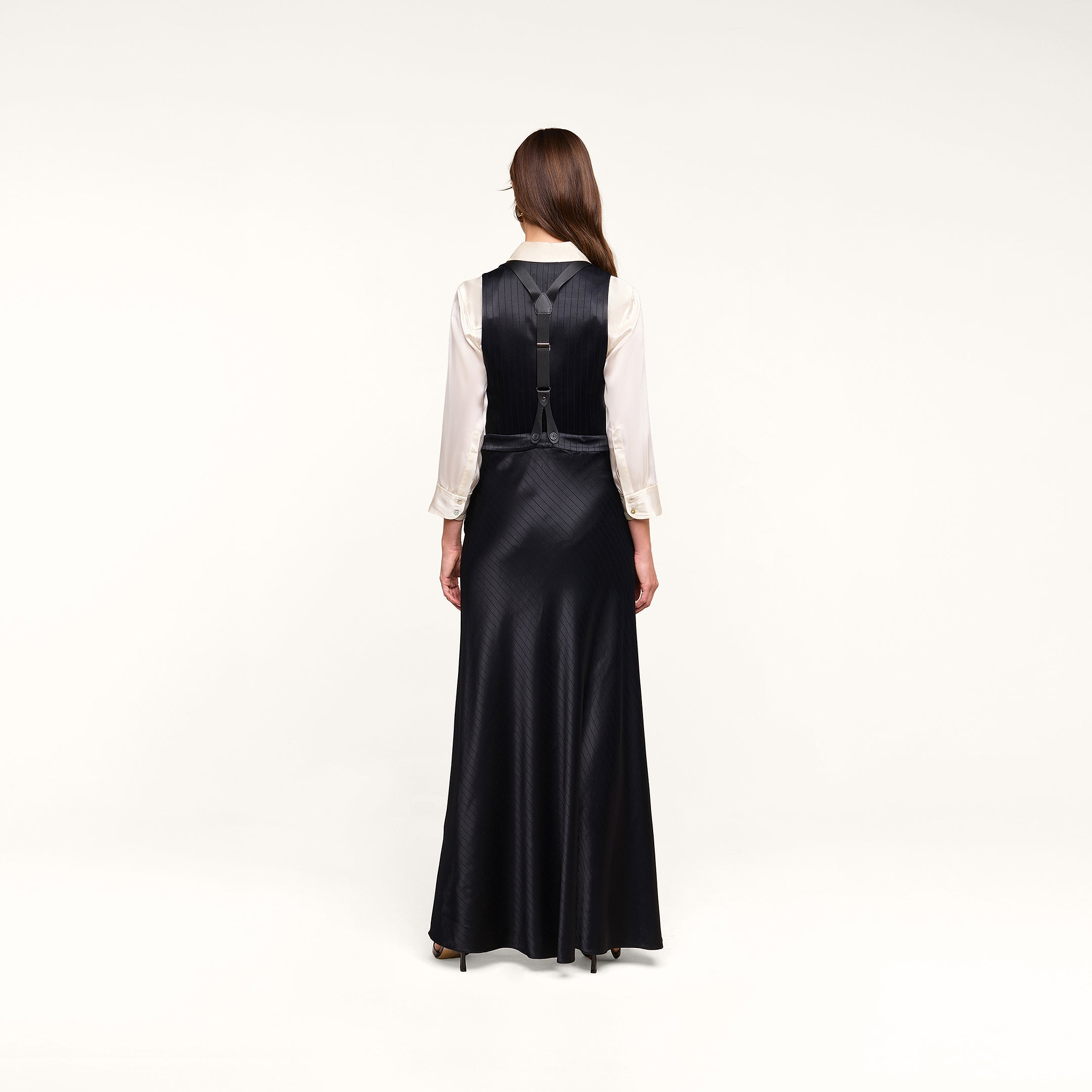 A woman with long brown hair stands facing away, wearing the Vespera Suspender Maxi Skirt in black satin over a white blouse. The background is plain and white.