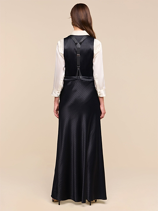A woman with long brown hair stands against a plain beige background, wearing a white blouse paired with the Vespera Suspender Maxi Skirt—a long black sleeveless skirt featuring adjustable suspenders and a harness detail on the back.