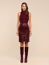 A woman stands facing forward against a plain white background, wearing the Violetta Vegan Leather Skirt—a croc-embossed vegan leather midi skirt—paired with a sleeveless burgundy top and coordinating high-heeled boots. She has long hair and minimal accessories.