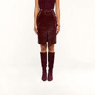 A person wearing a sleeveless burgundy top, the Violetta Vegan Leather Skirt featuring a croc-embossed texture and front slit, and matching burgundy knee-high boots stands against a white background.
