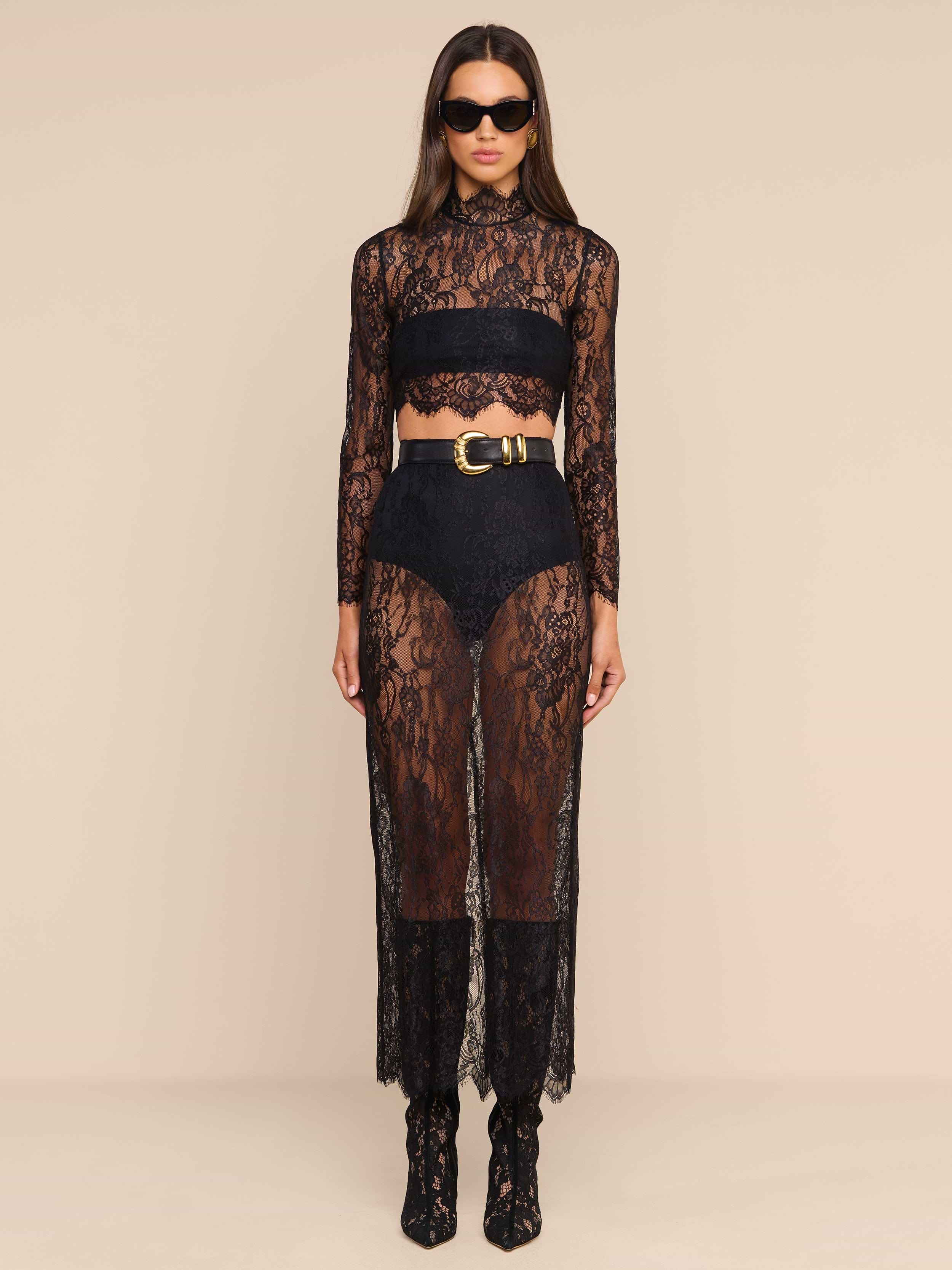 Woman wearing a black chantilly lace sheer long-sleeve crop top paired with the Raymonde Lace Maxi Skirt, standing against a beige background, accessorized with sunglasses and ankle boots—an effortlessly chic look perfect for a night out.