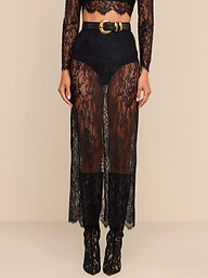 A person wearing high-waisted black shorts layered under a sheer black Raymonde Lace Maxi Skirt with a gold-buckled belt, paired with matching lace boots—perfect for a stylish night out. The top part of the body is mostly out of frame.