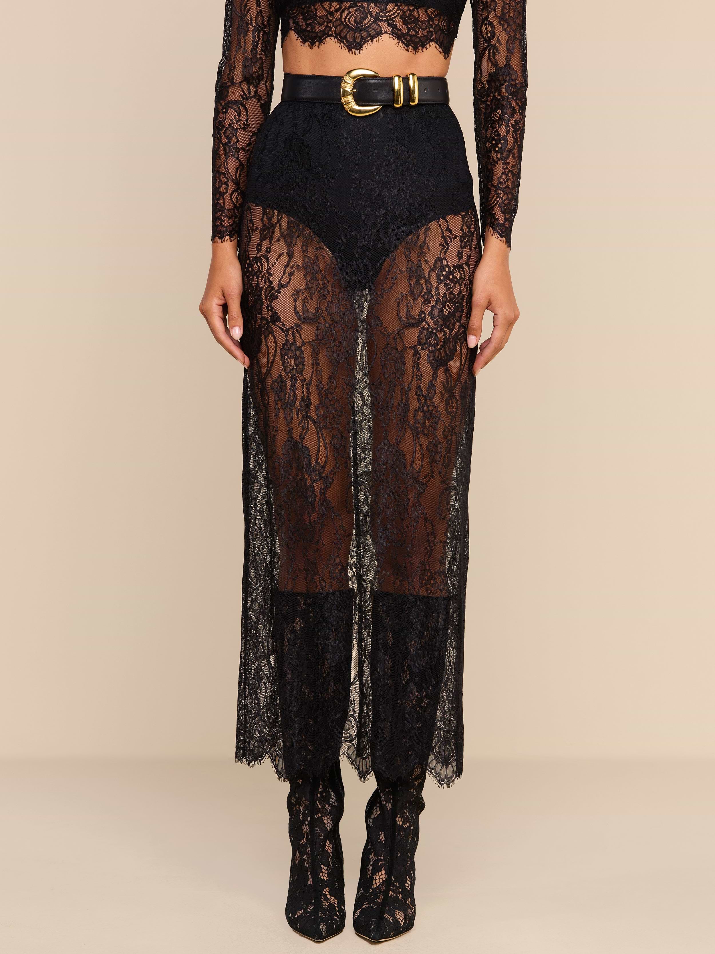 A person wearing high-waisted black shorts layered under a sheer black Raymonde Lace Maxi Skirt with a gold-buckled belt, paired with matching lace boots—perfect for a stylish night out. The top part of the body is mostly out of frame.