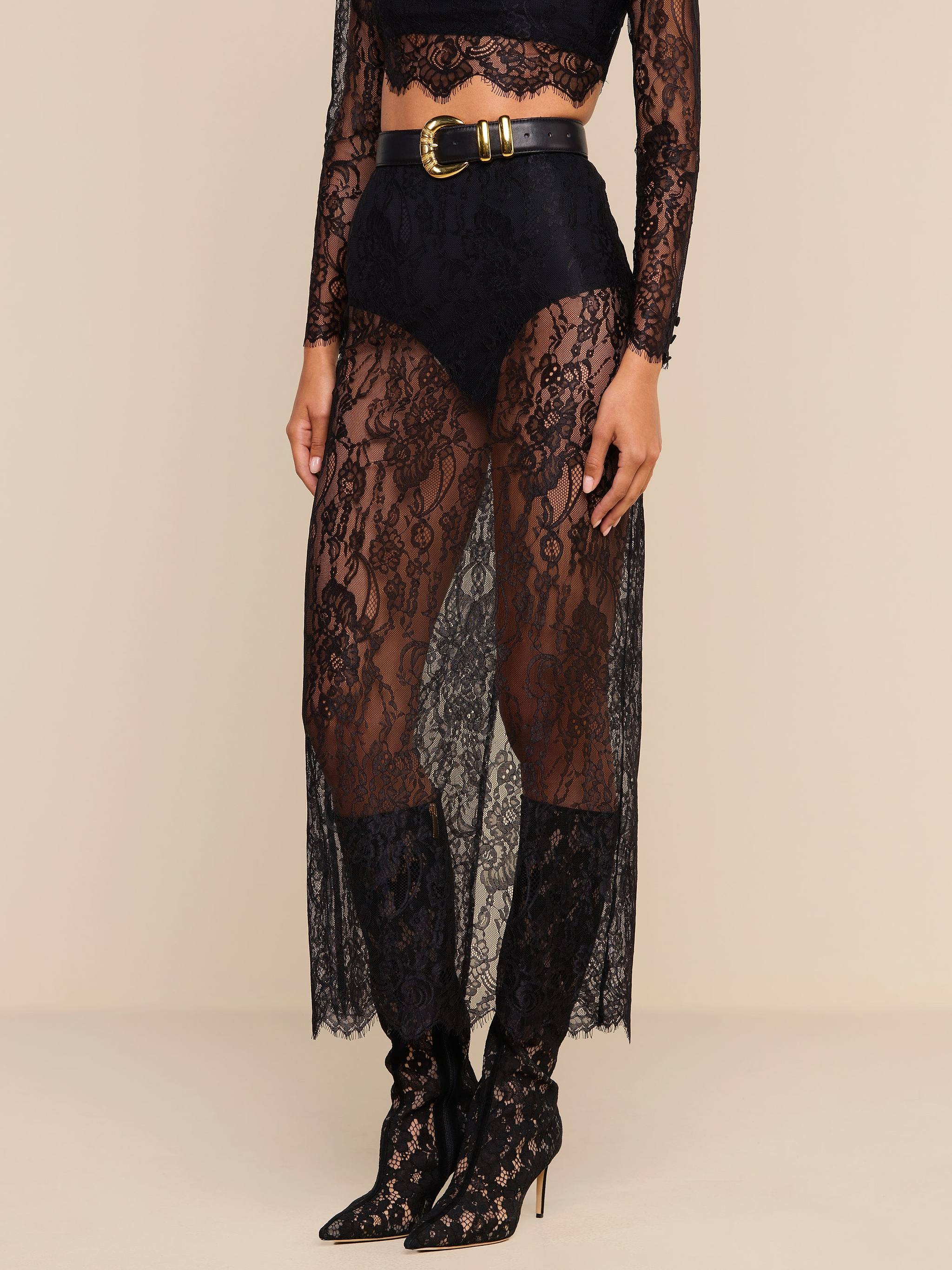 Woman wearing a sheer black Raymonde Lace Maxi Skirt with high-waisted black briefs underneath, a black lace long-sleeve crop top, and black pointed-toe, lace-up high heel boots—perfect for a stylish night out. A black belt with a gold buckle completes the look.