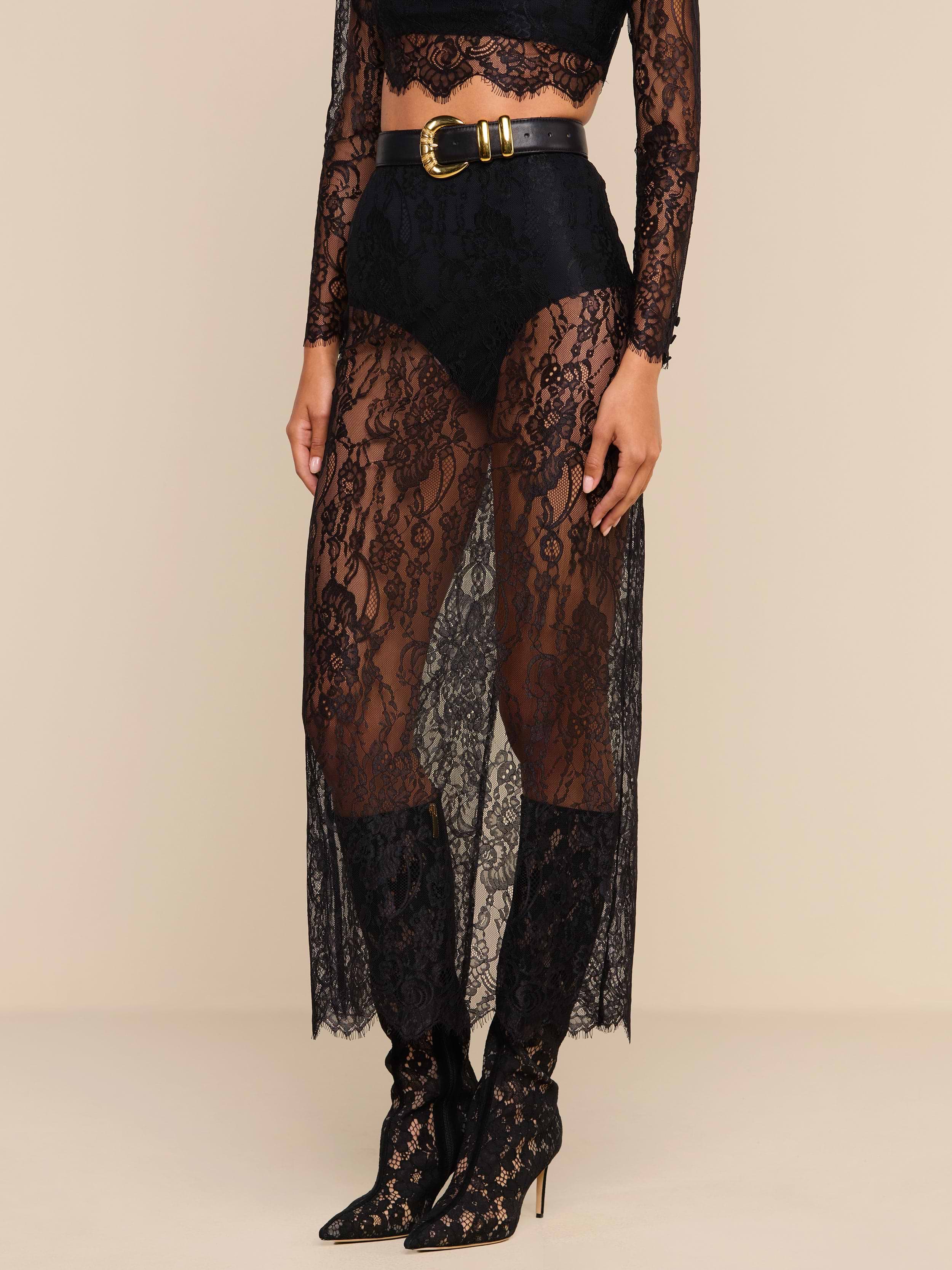 Woman wearing a sheer black Raymonde Lace Maxi Skirt with high-waisted black briefs underneath, a black lace long-sleeve crop top, and black pointed-toe, lace-up high heel boots—perfect for a stylish night out. A black belt with a gold buckle completes the look.