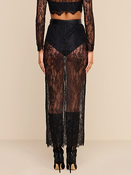 A person stands facing away, wearing the Raymonde Lace Maxi Skirt featuring a sheer black Chantilly lace with a floral pattern over black underwear and heeled shoes—an effortlessly chic look perfect for a night out. The background is plain beige.