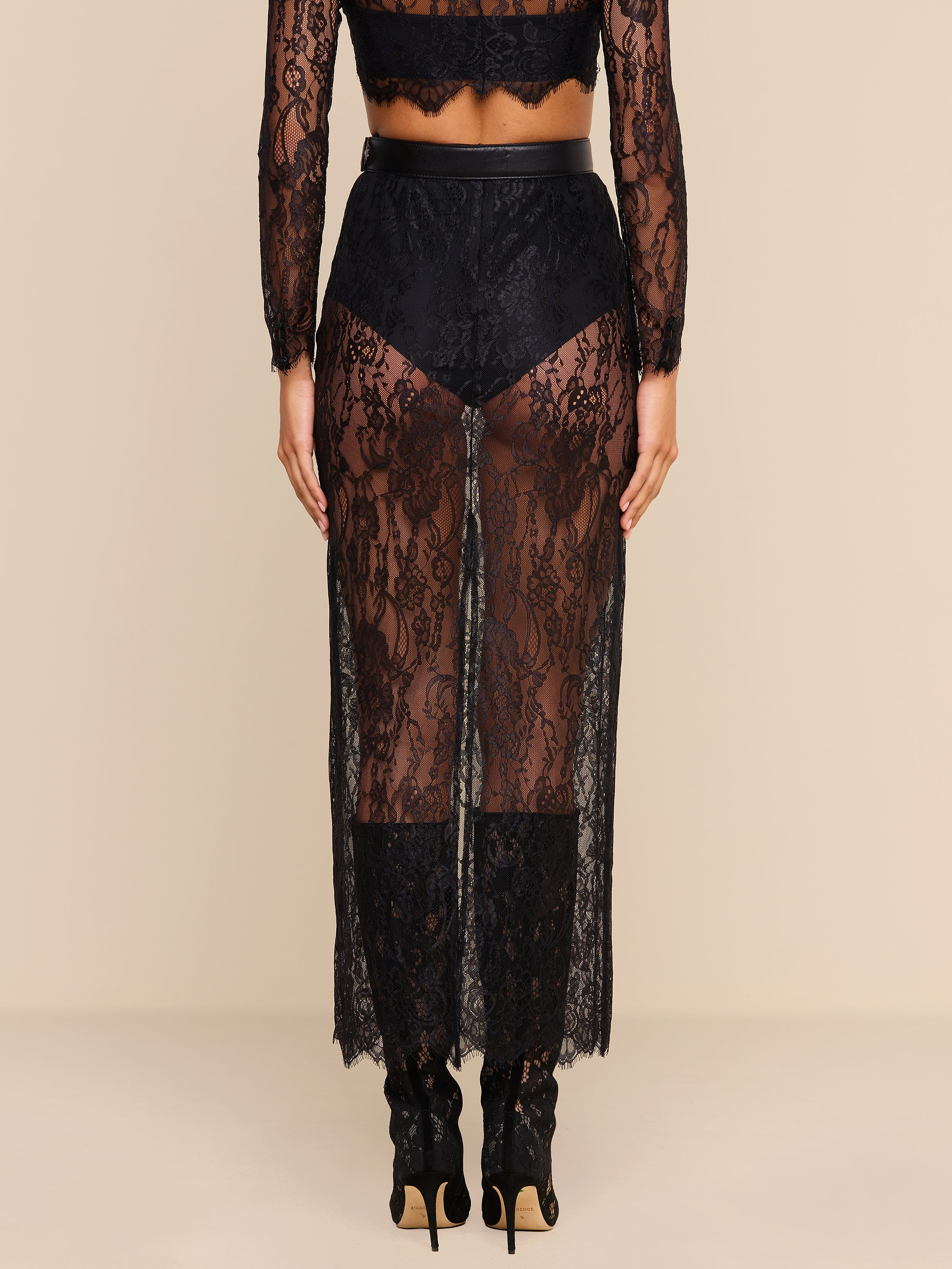 A person stands facing away, wearing the Raymonde Lace Maxi Skirt featuring a sheer black Chantilly lace with a floral pattern over black underwear and heeled shoes—an effortlessly chic look perfect for a night out. The background is plain beige.