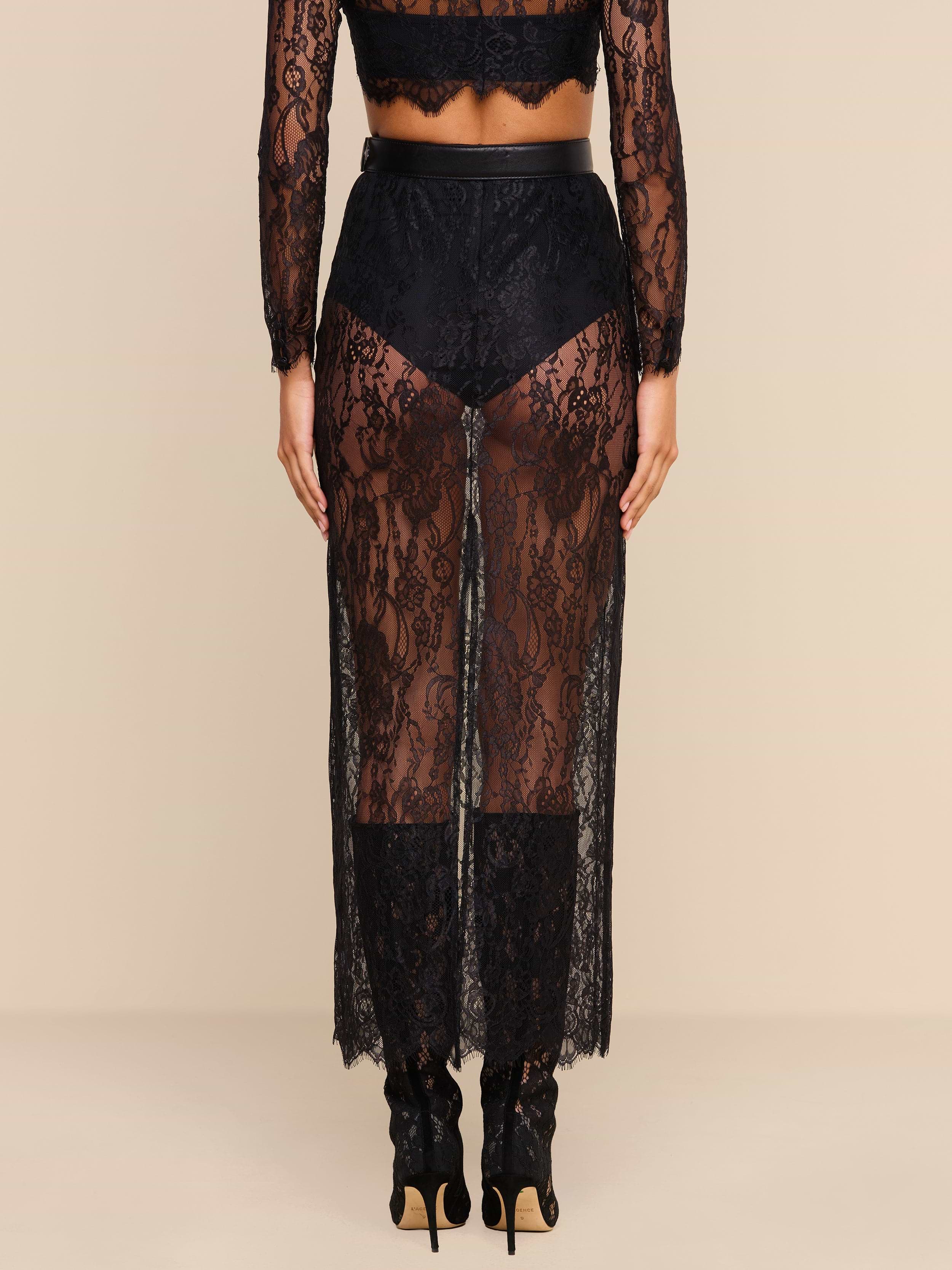 A person stands facing away, wearing the Raymonde Lace Maxi Skirt featuring a sheer black Chantilly lace with a floral pattern over black underwear and heeled shoes—an effortlessly chic look perfect for a night out. The background is plain beige.