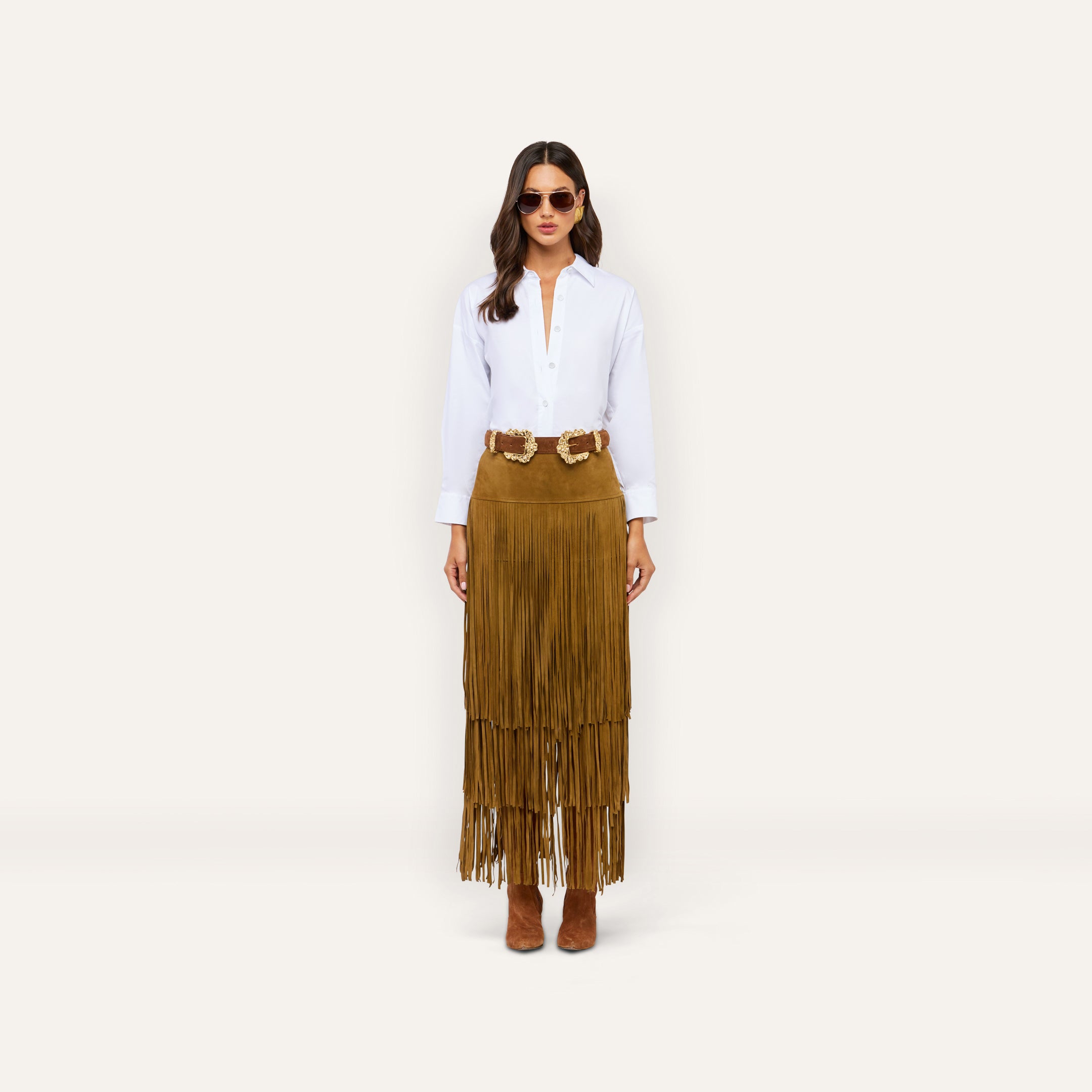 A person wearing a white button-down shirt, a double-buckle belt, and a Karoline Suede Fringe Skirt, which is a long brown tiered fringe statement piece.