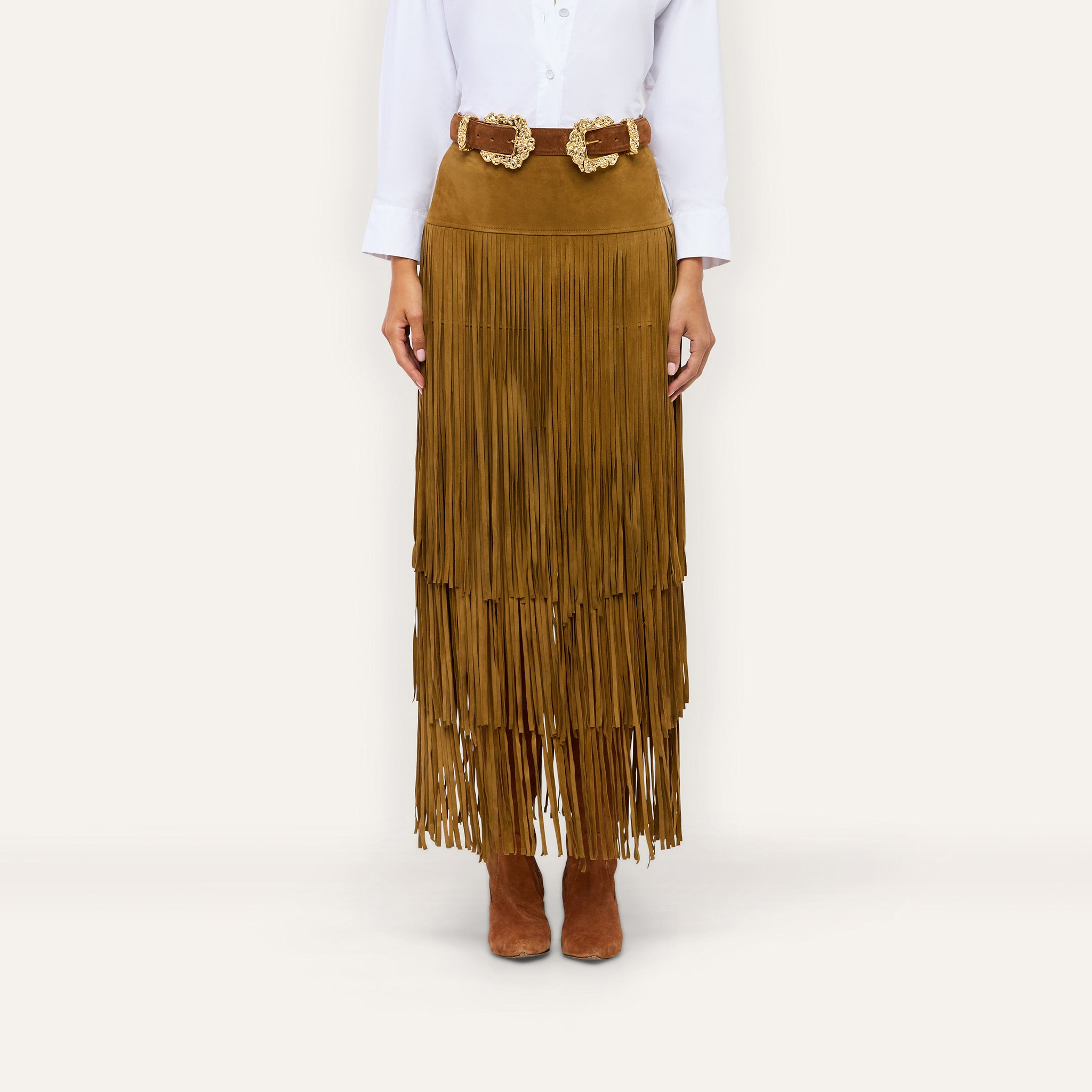 The Karoline Suede Fringe Skirt, a brown tiered fringe design, is paired with a double-buckle belt, white button-down shirt, and brown ankle boots for a stylish ensemble.