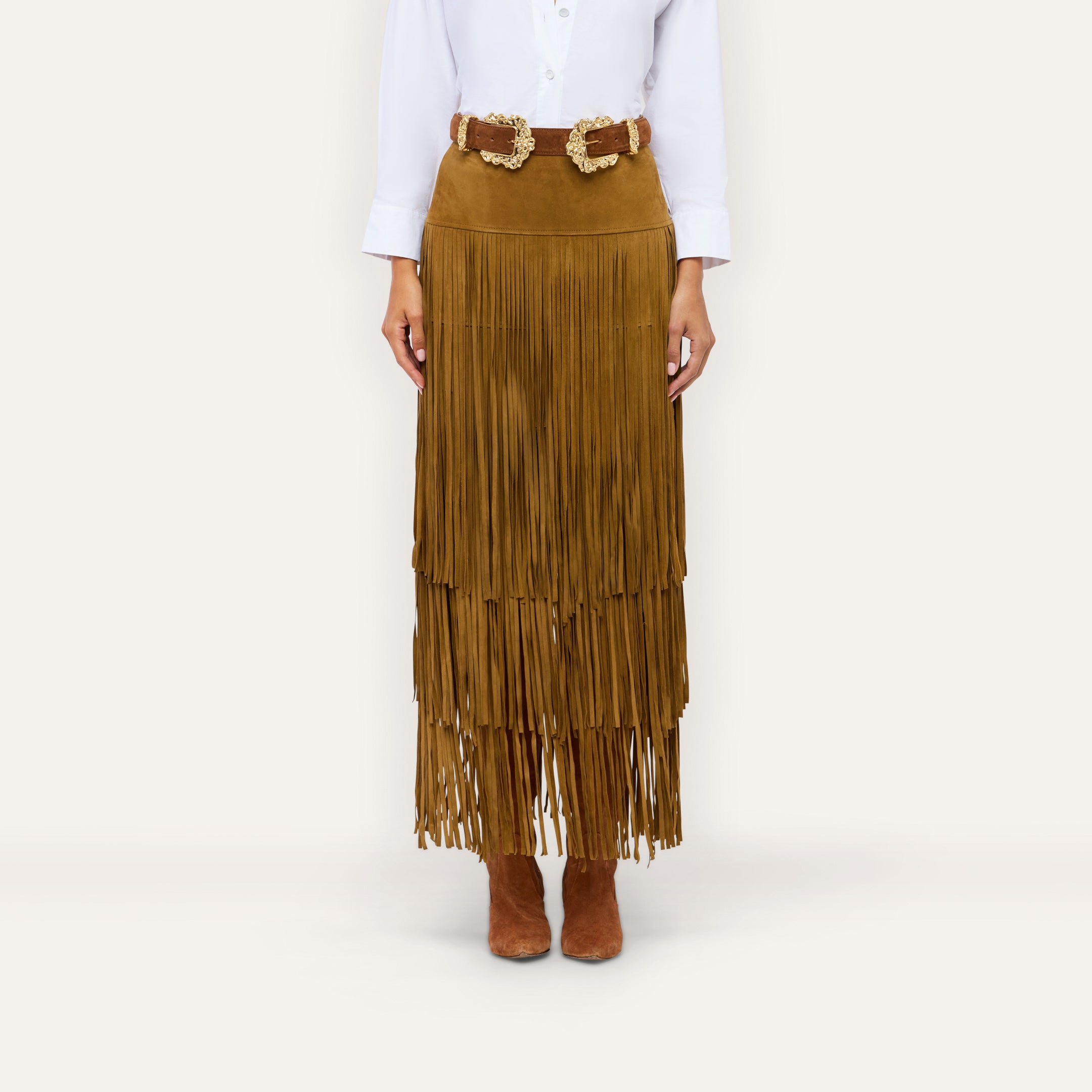 The Karoline Suede Fringe Skirt, a brown tiered fringe design, is paired with a double-buckle belt, white button-down shirt, and brown ankle boots for a stylish ensemble.