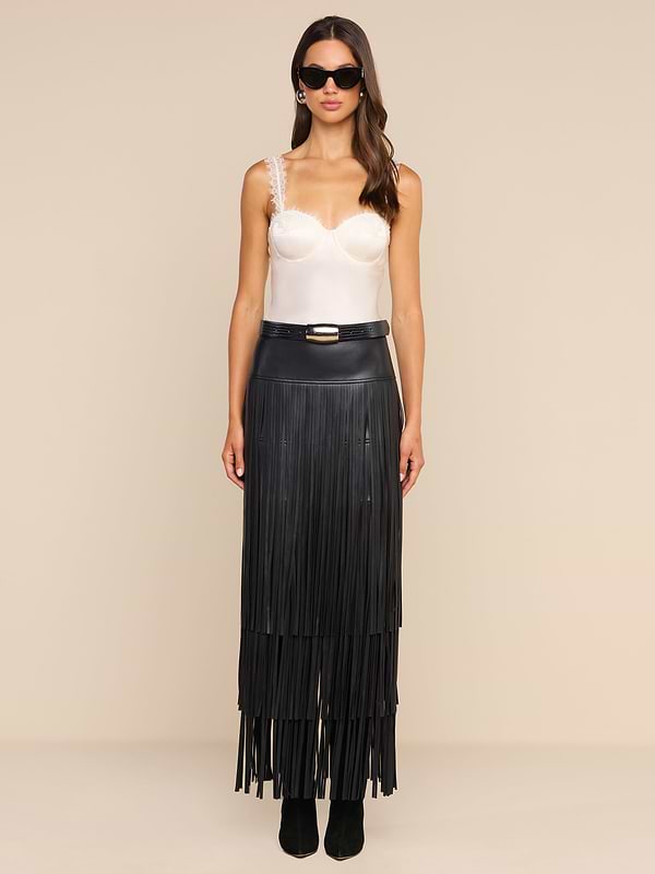 A woman stands against a plain beige background, facing forward with a neutral expression. She wears sunglasses, a white satin camisole with lace straps, and the high-waisted Karoline Fringe Skirt in black vegan leather, featuring long fringe details.
