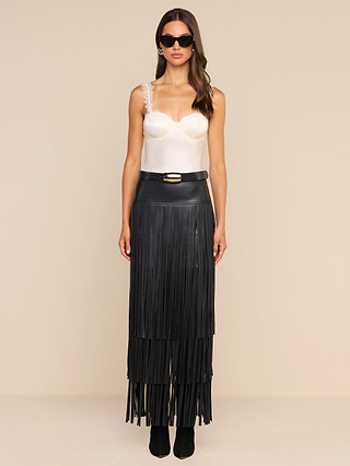 A woman stands against a plain beige background, facing forward with a neutral expression. She wears sunglasses, a white satin camisole with lace straps, and the high-waisted Karoline Fringe Skirt in black vegan leather, featuring long fringe details.