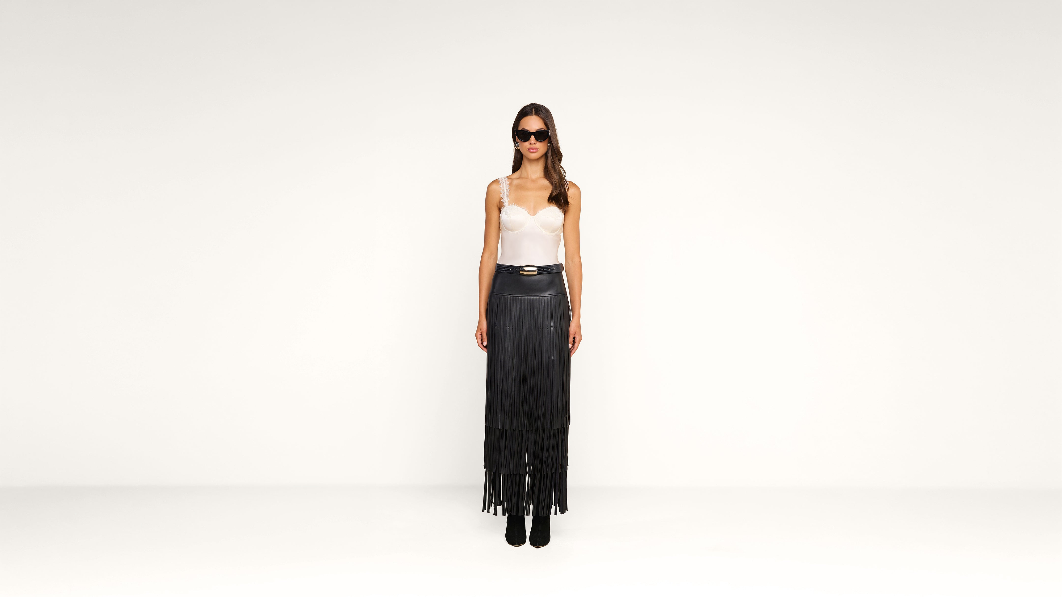 Woman stands against a plain white background, wearing sunglasses, a white top, a black high-waisted skirt with fringe, and black ankle boots. She faces forward with her arms relaxed at her sides.