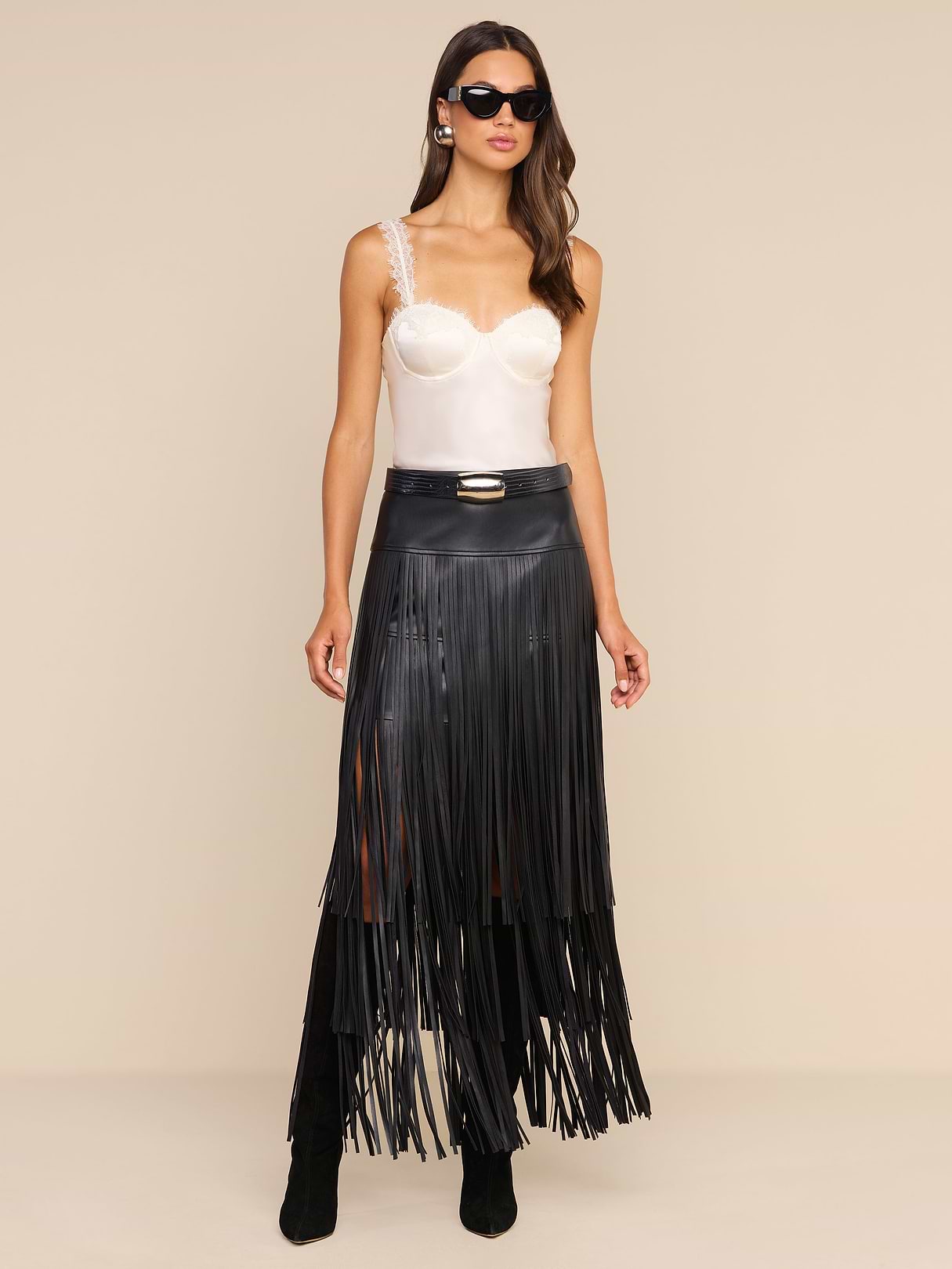 A woman wearing black sunglasses, a white satin camisole, and the Karoline Fringe Skirt with black boots stands against a beige background.