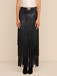 Wearing a white sleeveless top, a person models the Karoline Fringe Skirt—a long, black, tiered fringe maxi skirt with a belt and buckle—while standing against a plain beige background.
