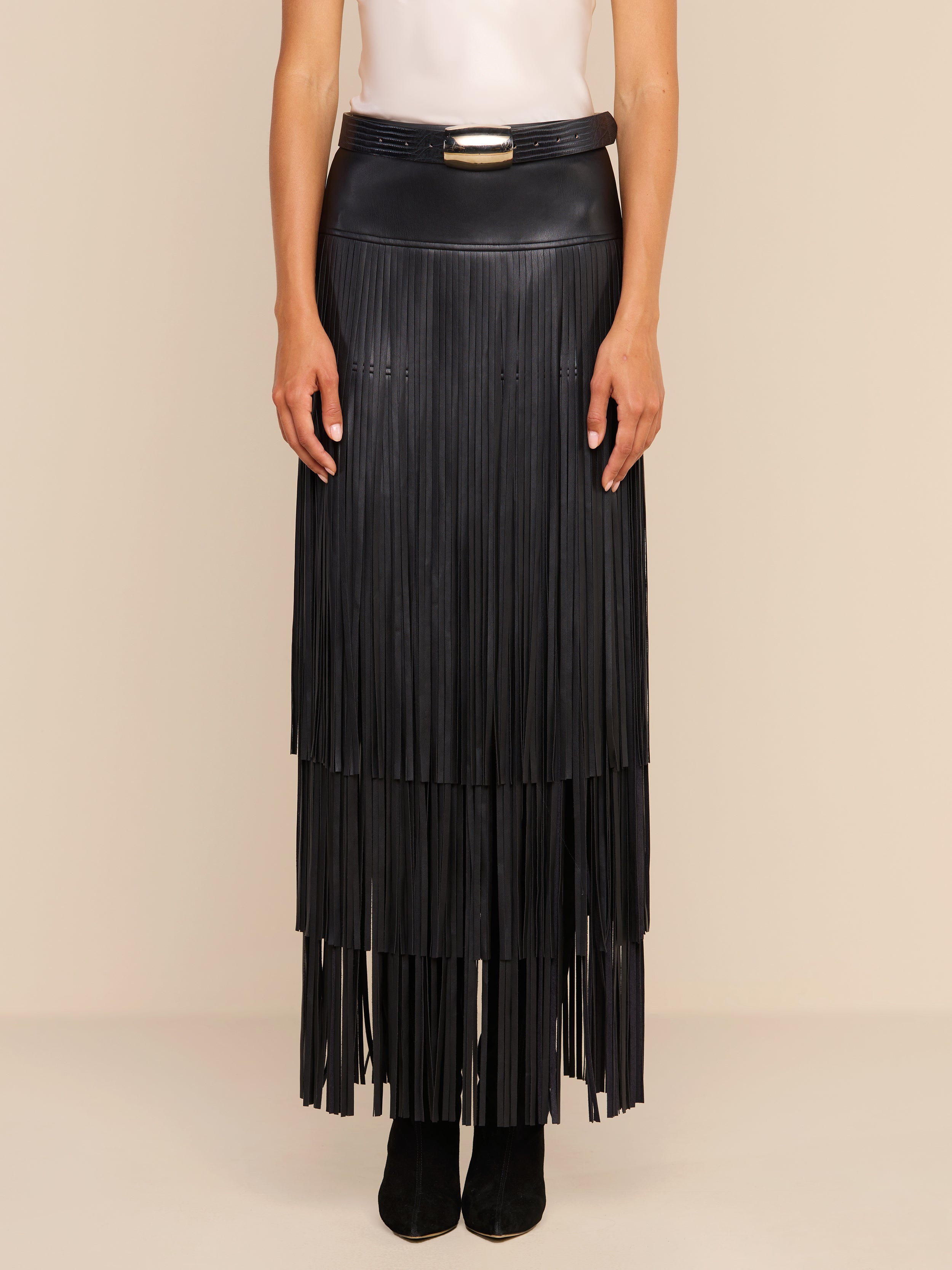 Wearing a white sleeveless top, a person models the Karoline Fringe Skirt—a long, black, tiered fringe maxi skirt with a belt and buckle—while standing against a plain beige background.
