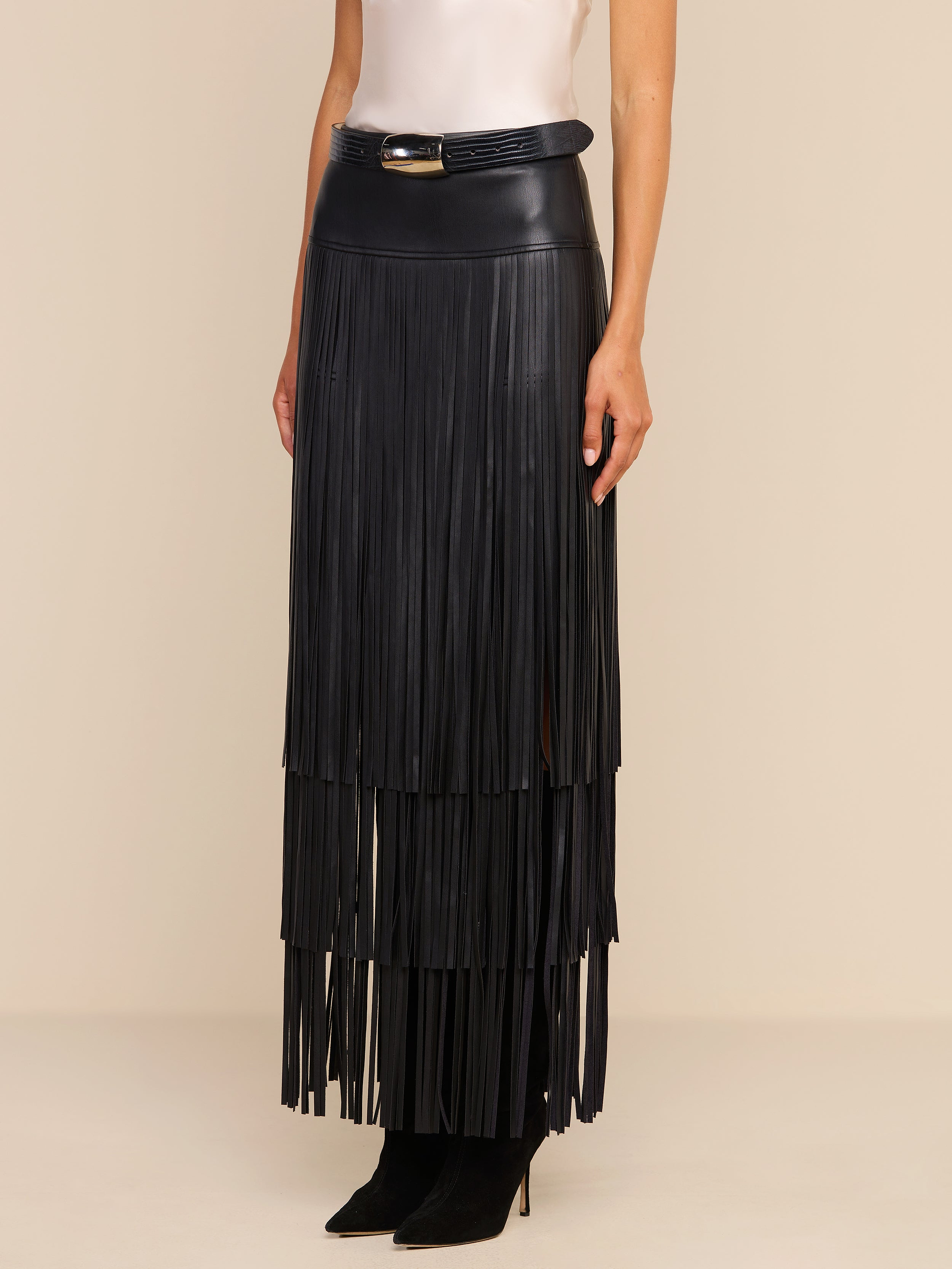 A woman wears the Karoline Fringe Skirt—a high-waisted, black vegan leather maxi skirt with long fringe layers—paired with a belt featuring a gold buckle, a white sleeveless top, and black pointed-toe boots. Only her lower body is visible.