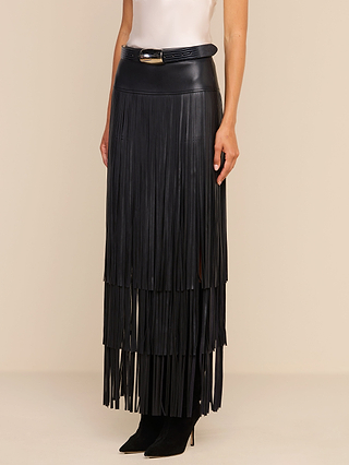A woman wears the Karoline Fringe Skirt—a high-waisted, black vegan leather maxi skirt with long fringe layers—paired with a belt featuring a gold buckle, a white sleeveless top, and black pointed-toe boots. Only her lower body is visible.