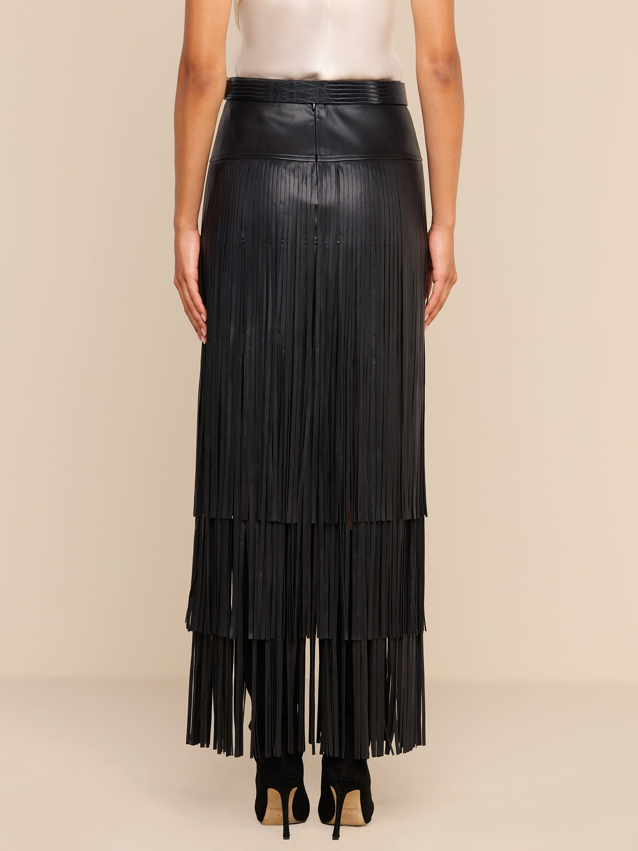 A person stands with their back to the camera, wearing a sleeveless light top and the Karoline Fringe Skirt—a long black vegan leather fringe maxi skirt—paired with high heels, against a beige background.