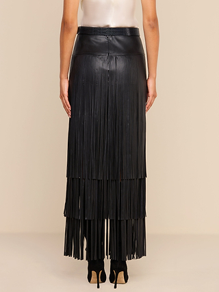 A person stands with their back to the camera, wearing a sleeveless light top and the Karoline Fringe Skirt—a long black vegan leather fringe maxi skirt—paired with high heels, against a beige background.