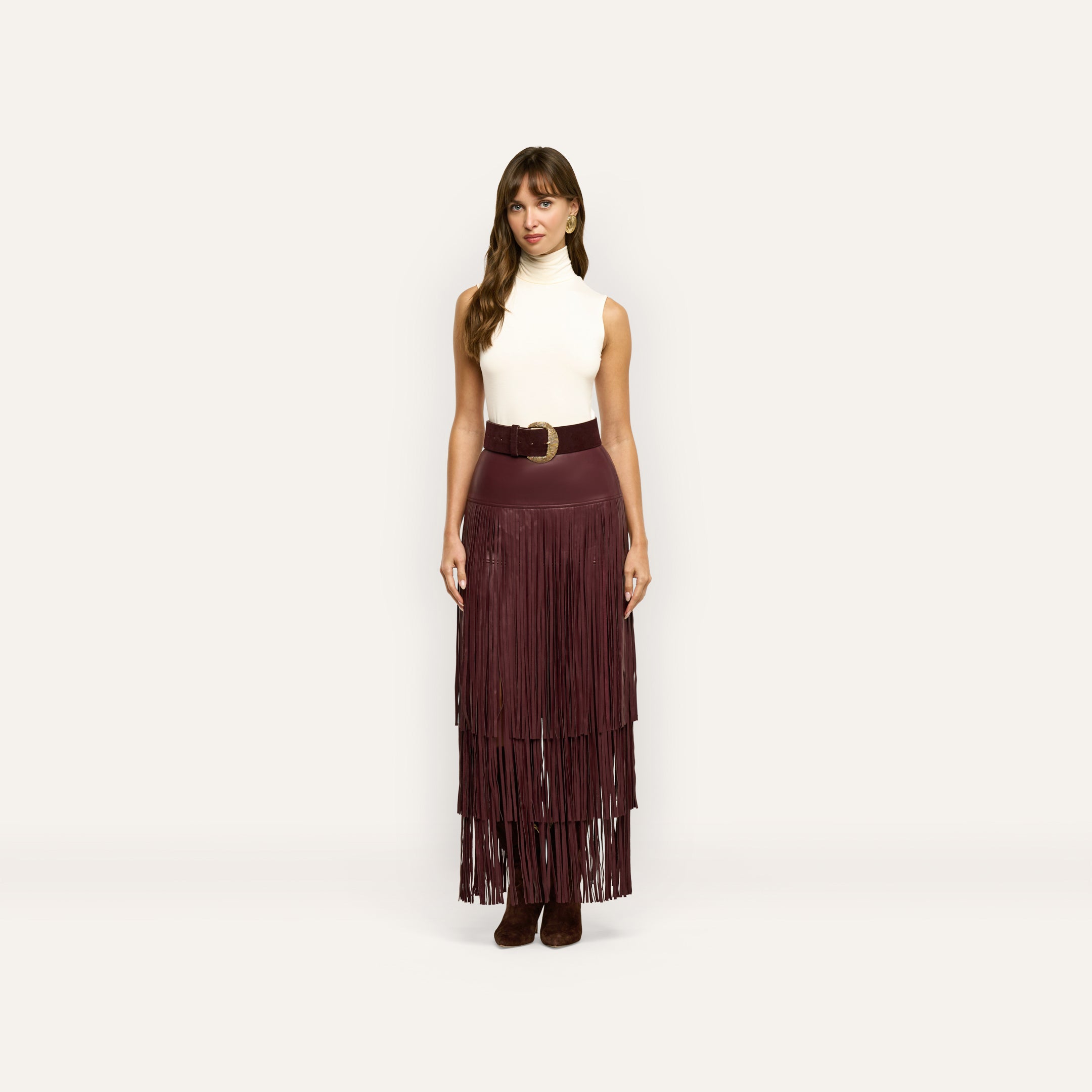 A woman wearing a sleeveless white turtleneck and a long, tiered burgundy Karoline Fringe Skirt cinched with a wide belt.