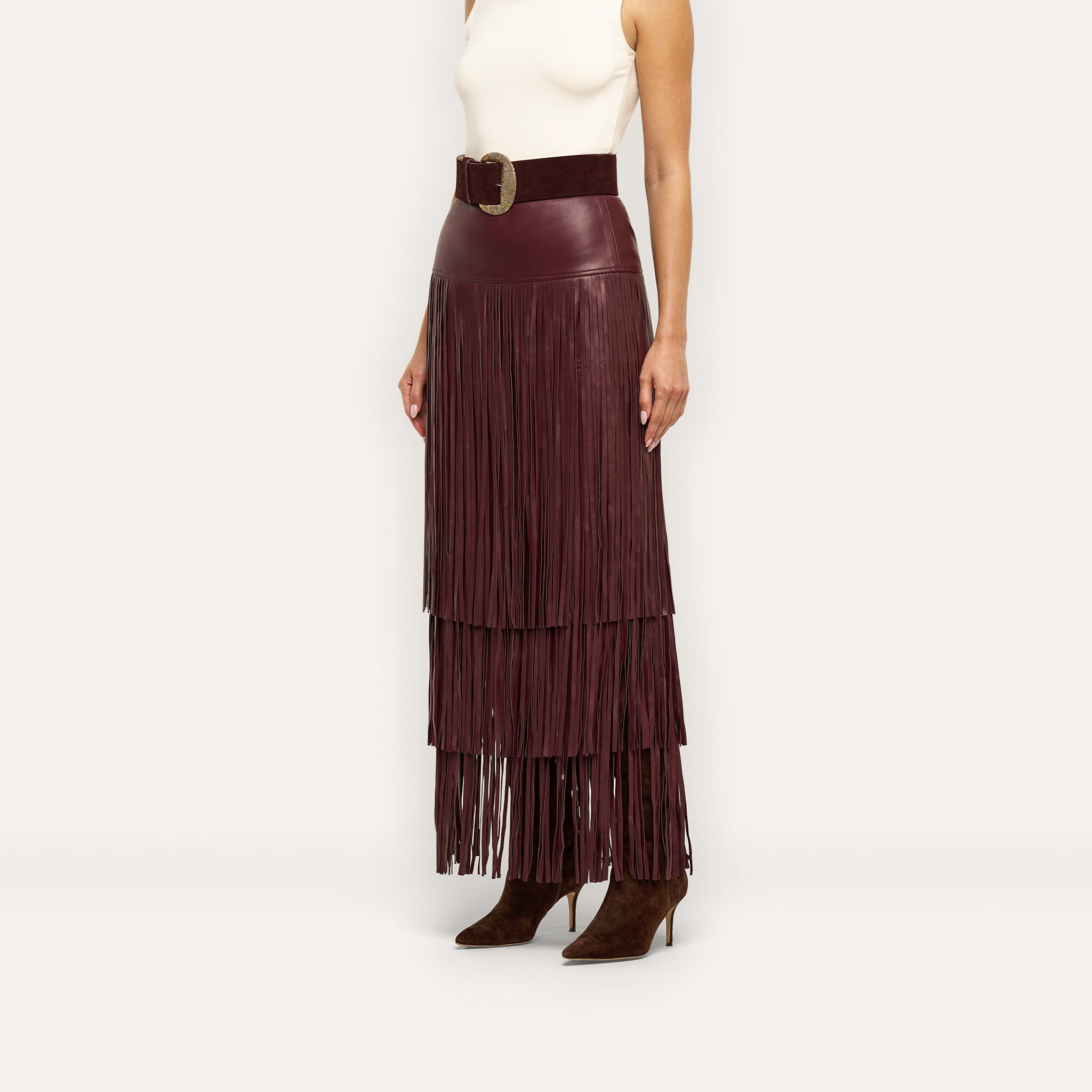 A person wears a burgundy Karoline Fringe Skirt featuring layered fringe, a wide matching belt, and brown suede heeled boots, styled as a deep red vegan leather statement piece.