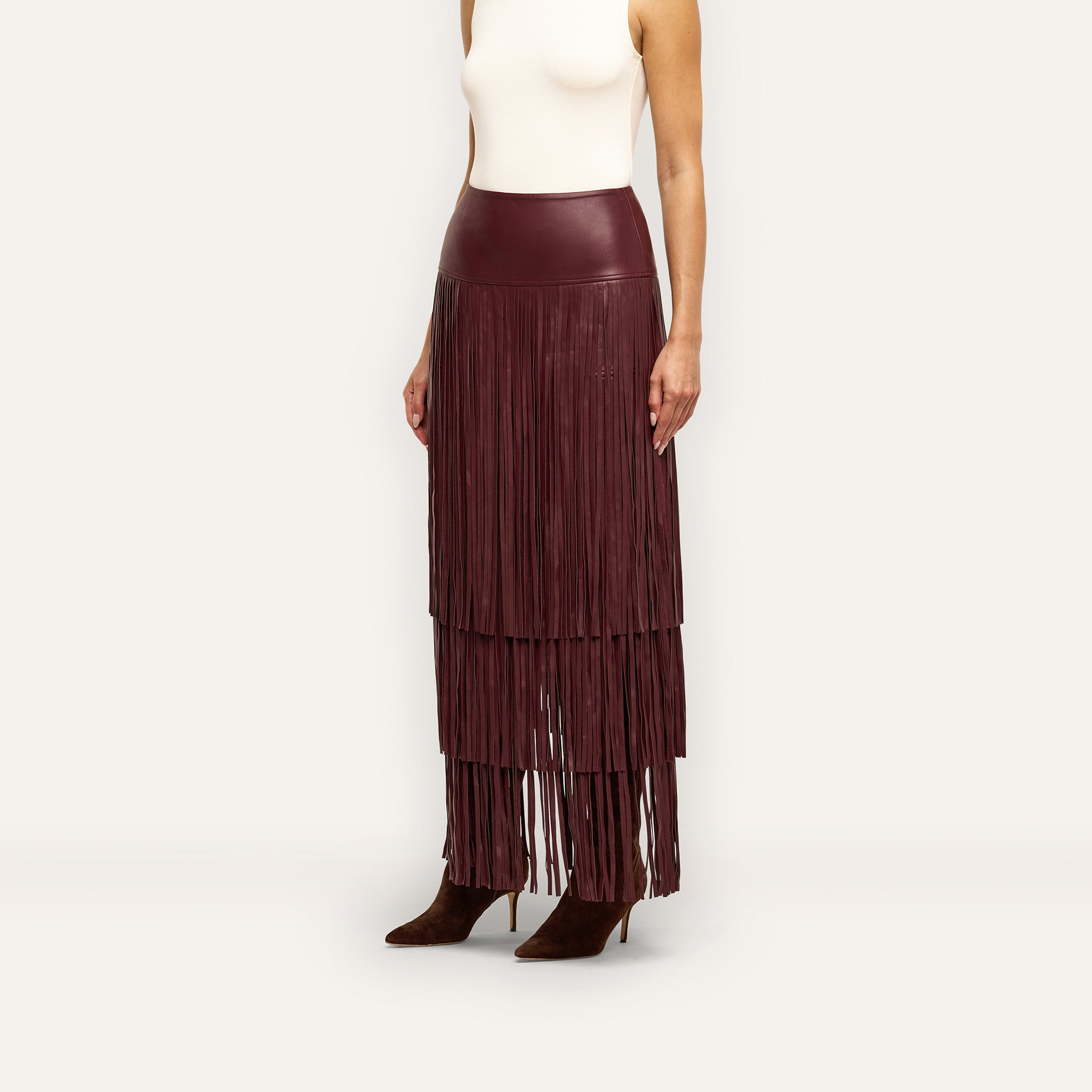 A Karoline Fringe Skirt in high-waisted, deep red vegan leather worn with a white sleeveless top and brown pointed-toe heels.