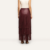 A person wearing a floor-length, deep red vegan leather Karoline Fringe Skirt featuring three tiers of fringe.