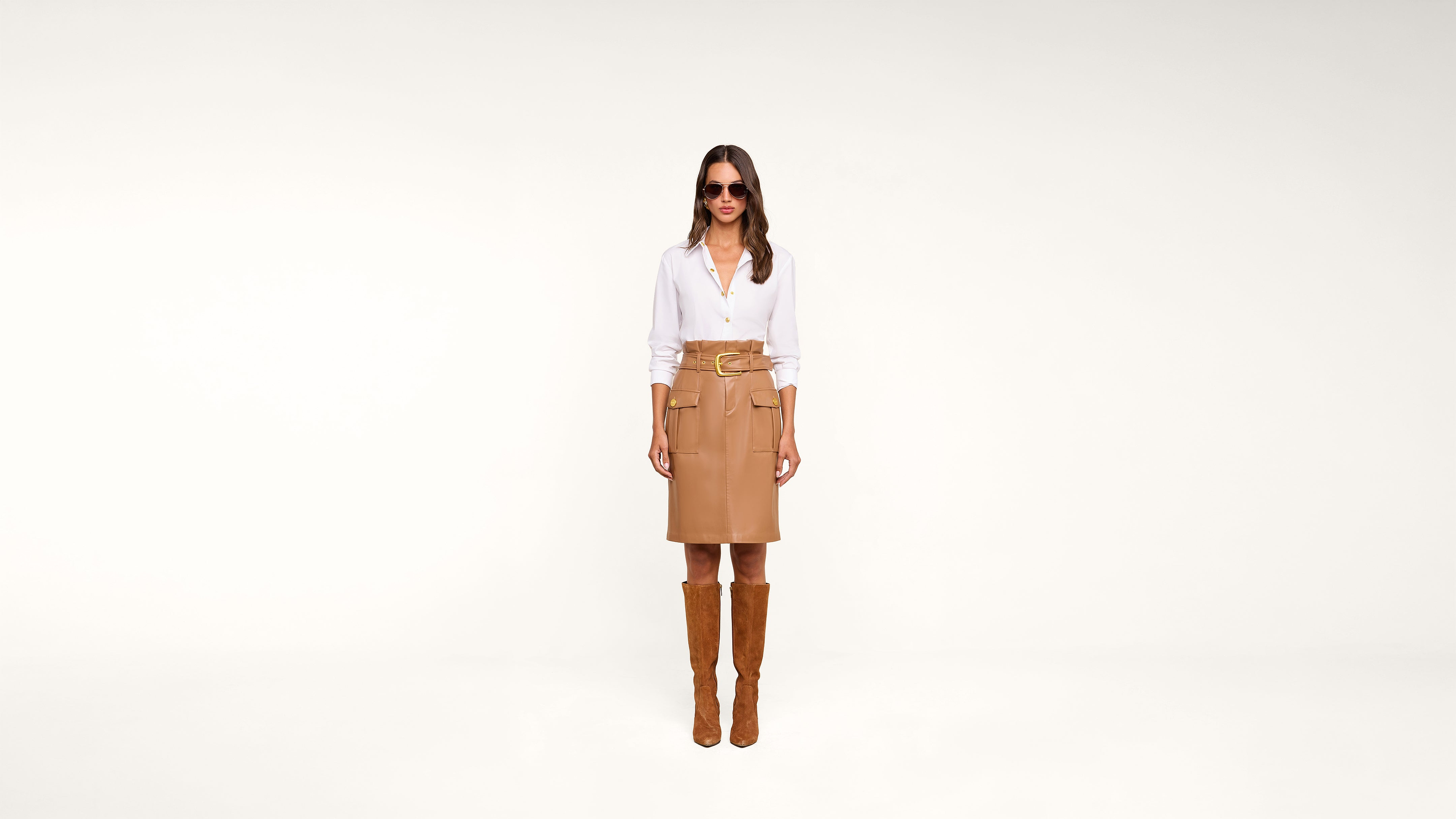 Woman stands against a plain white background wearing sunglasses, a white blouse, a tan belted skirt, and tall brown boots.