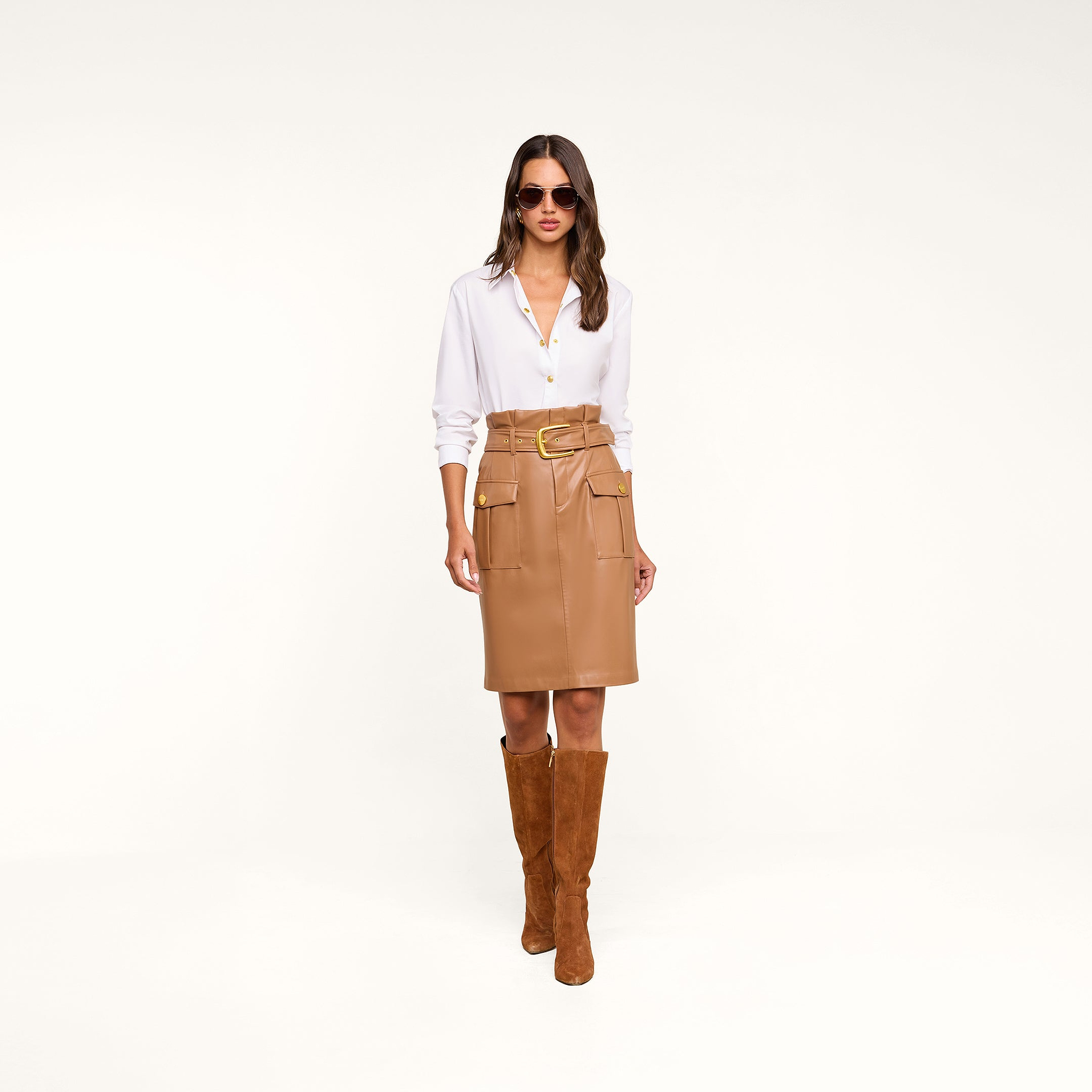A woman wearing sunglasses, a white button-down shirt, the Kaelis Vegan Leather Skirt with pockets and a paperbag waist in cognac, and brown suede knee-high boots stands against a beige background.