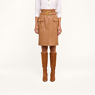 A person wears a white button-down shirt, the Kaelis Vegan Leather Skirt in tan with a paperbag waist, large pockets, and gold buttons, paired with knee-high brown suede boots, standing against a beige background.