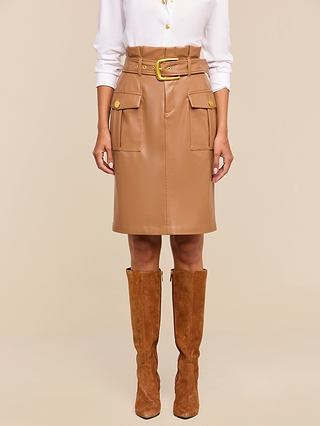 A person wears a white button-down shirt, the Kaelis Vegan Leather Skirt in tan with a paperbag waist, large pockets, and gold buttons, paired with knee-high brown suede boots, standing against a beige background.
