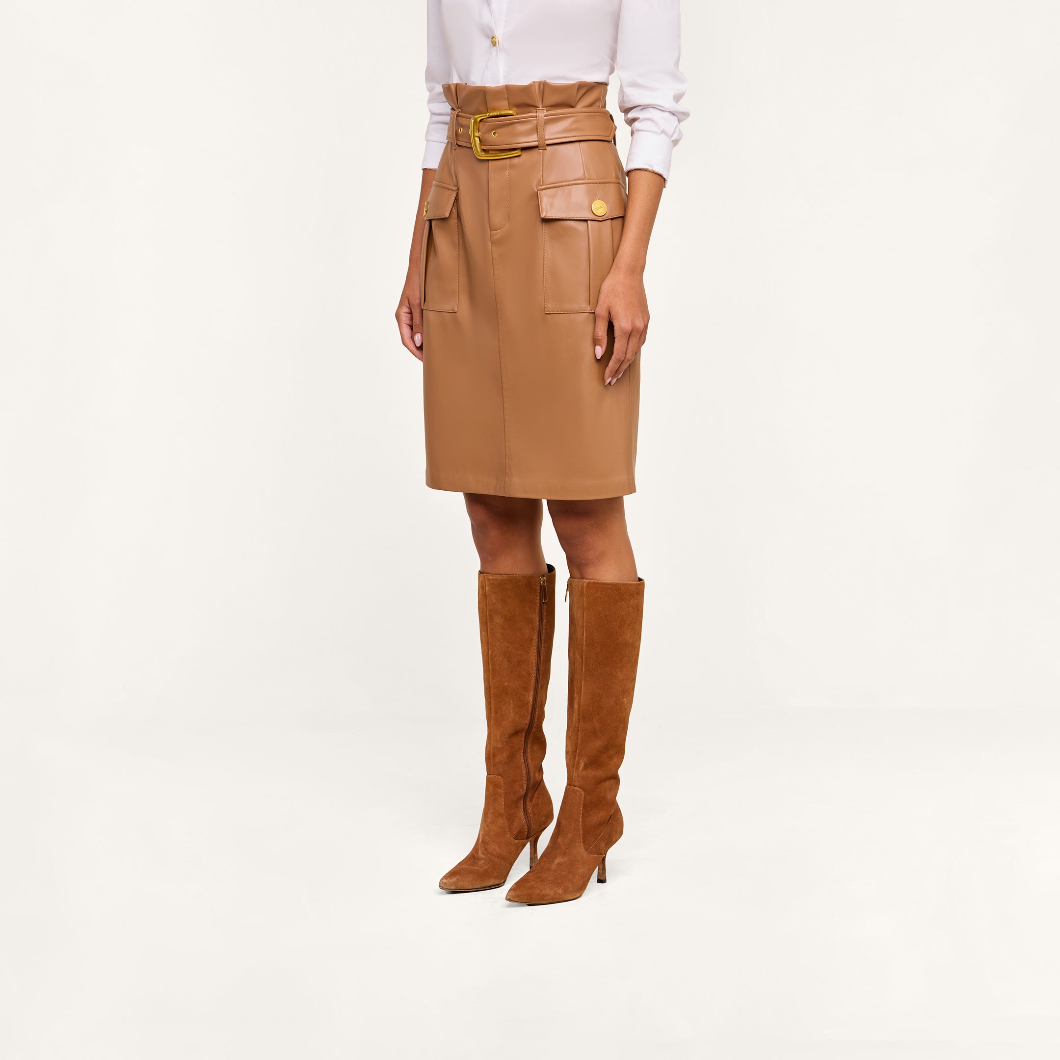 A woman stands against a beige background wearing a white long-sleeve shirt, the Kaelis Vegan Leather Skirt in tan with a paperbag waist, large pockets, and gold buttons, paired with brown suede knee-high heeled boots.