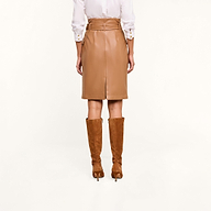 A person wearing a white shirt, the Kaelis Vegan Leather Skirt in cognac with a belt, and brown suede knee-high heeled boots is shown from the back against a neutral background.