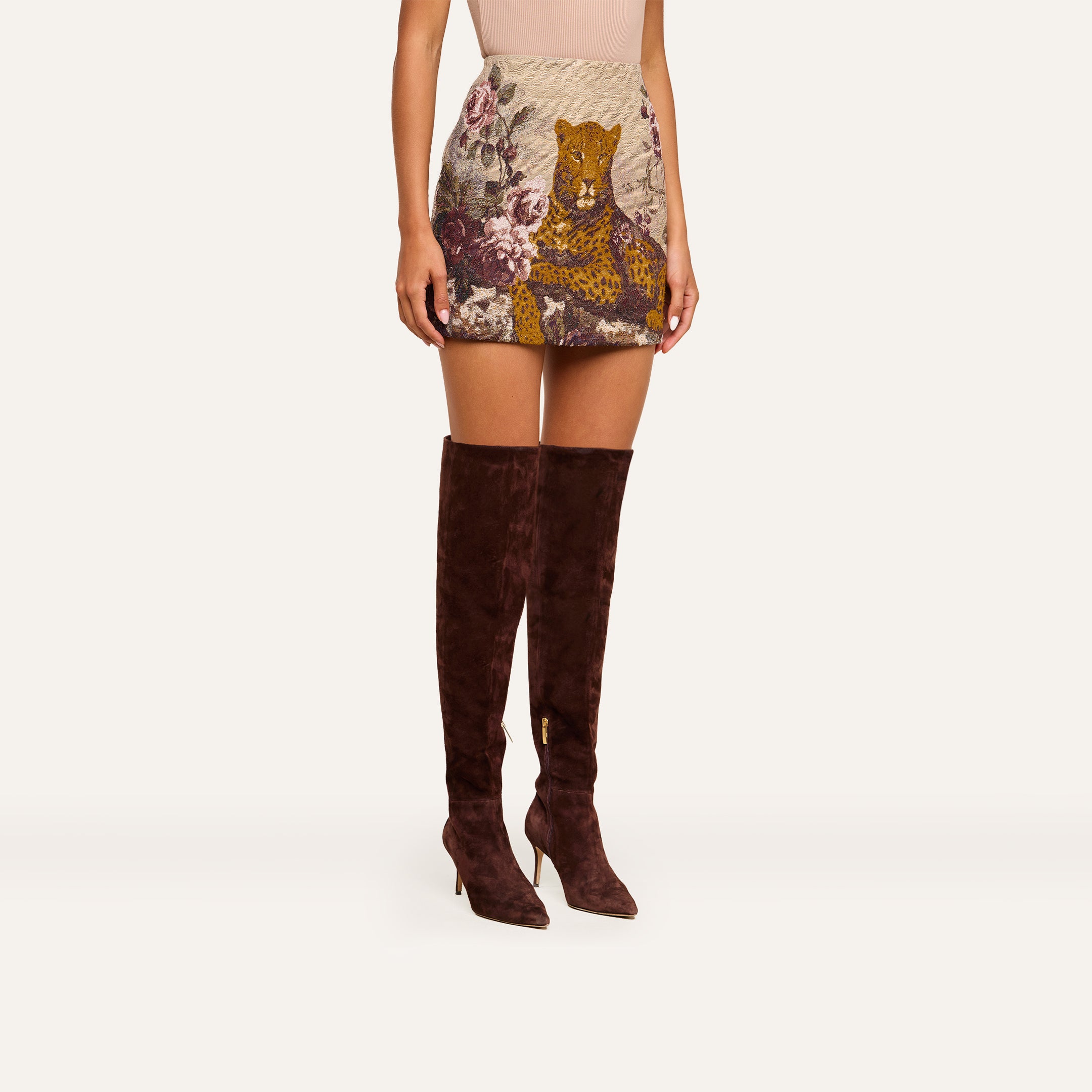 Dressed in the Marseille Mini Skirt, which features a vintage-inspired leopard and floral print, a woman styles her look with dark brown knee-high heeled boots.