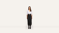 A woman stands facing forward, dressed in a white blouse, wide black belt, black tiered skirt, and black boots against a plain white background.