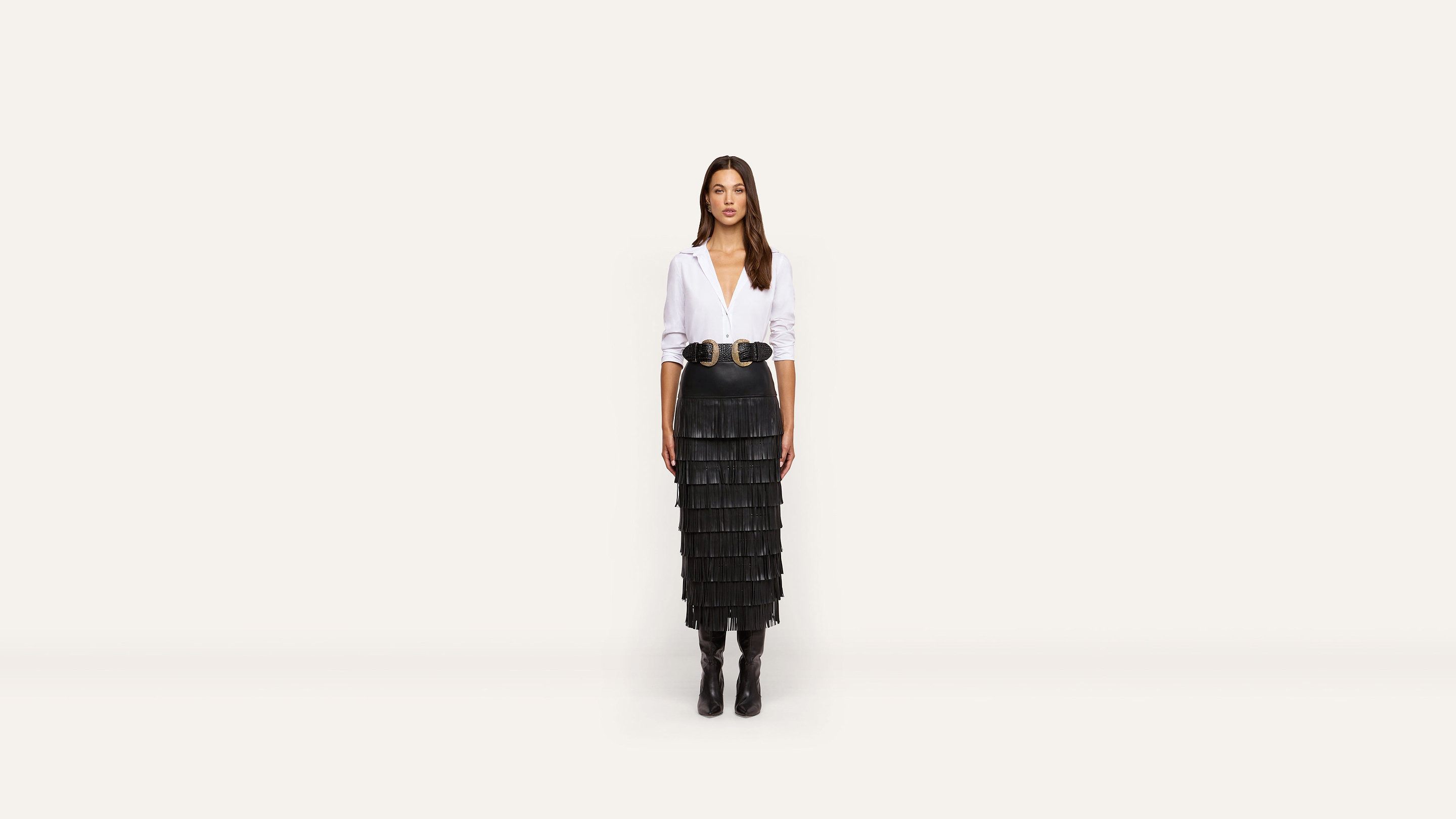 A woman stands facing forward, dressed in a white blouse, wide black belt, black tiered skirt, and black boots against a plain white background.
