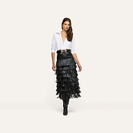 A woman models the Sevigne Fringe Skirt, a black vegan leather midi skirt with multiple tiers of long fringe, paired with a white button-down shirt, a wide double-buckle belt, and black boots.