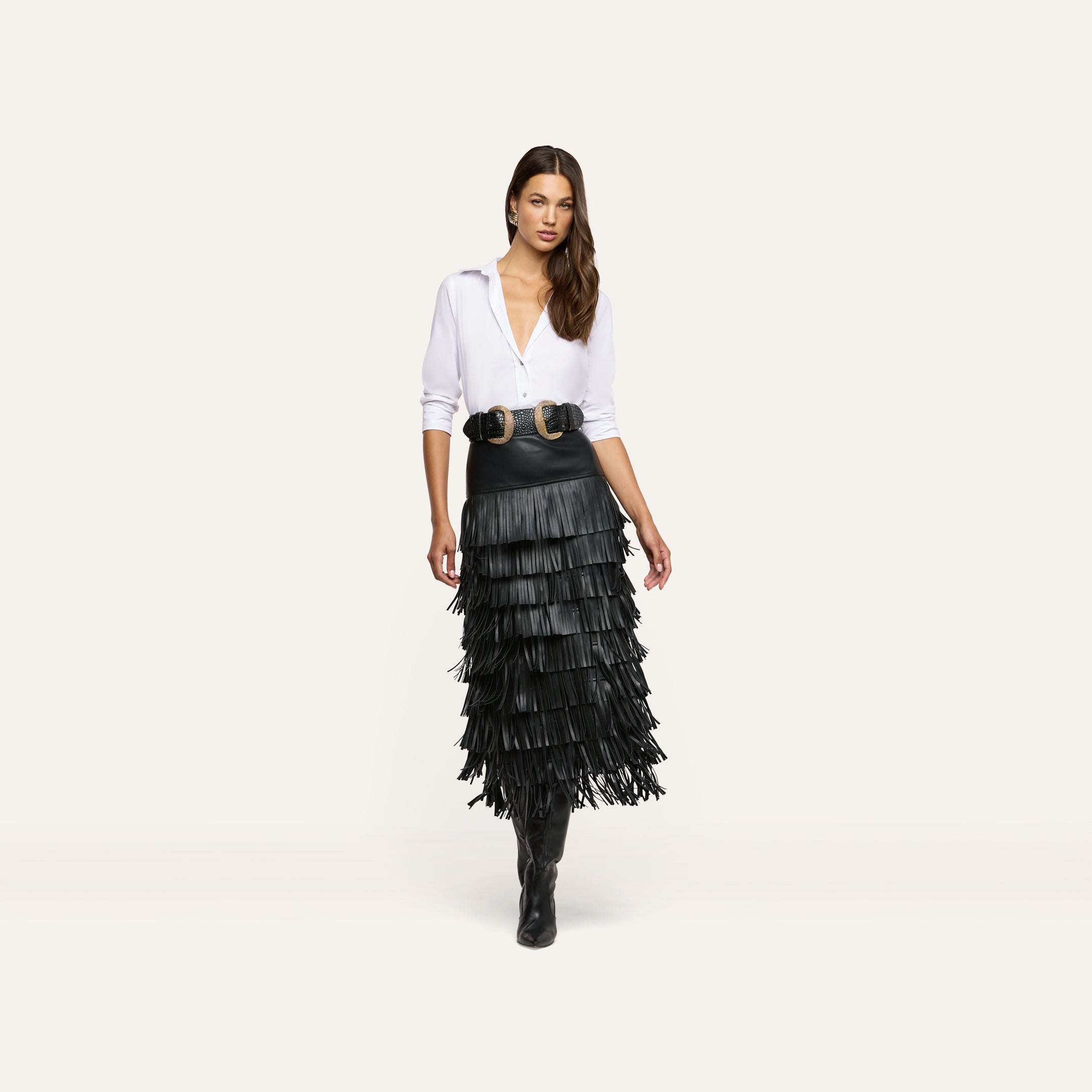 A woman models the Sevigne Fringe Skirt, a black vegan leather midi skirt with multiple tiers of long fringe, paired with a white button-down shirt, a wide double-buckle belt, and black boots.