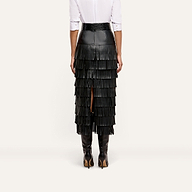 Rear view of a person wearing a Sevigne Fringe Skirt, featuring a high-waisted black midi design with multiple tiers of fringe and black knee-high boots.