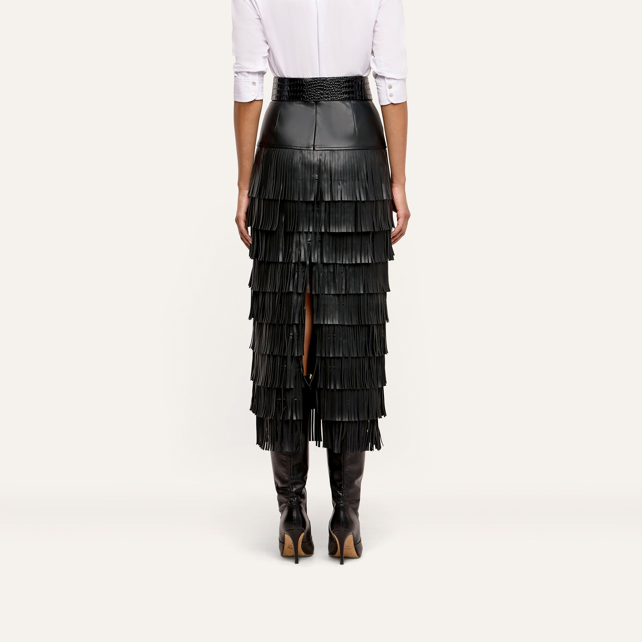 Rear view of a person wearing a Sevigne Fringe Skirt, featuring a high-waisted black midi design with multiple tiers of fringe and black knee-high boots.