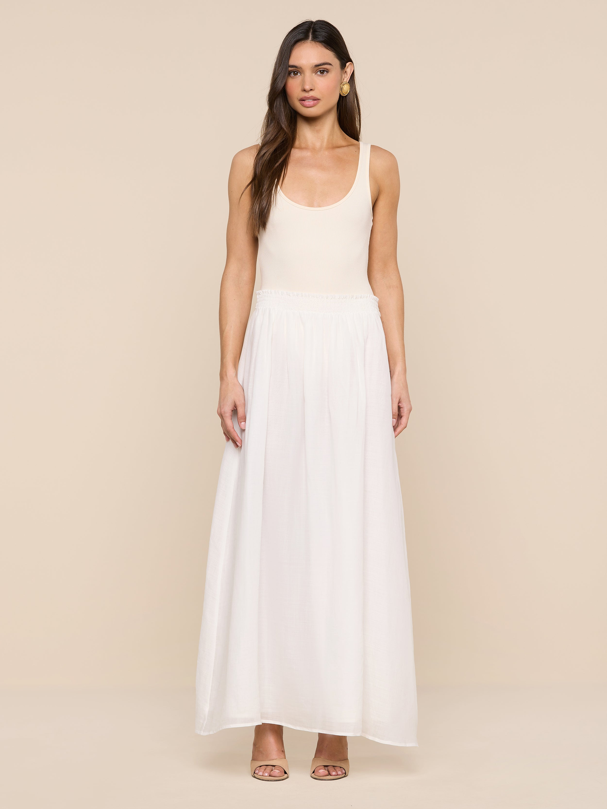A woman stands facing forward against a plain white background, wearing a sleeveless, light beige top paired with the Evangelina Maxi Skirt in white and heeled sandals. The breathable fabric of the Evangelina Maxi Skirt enhances her effortlessly elegant look.
