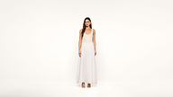 A woman stands facing forward, wearing a sleeveless, floor-length white dress and nude heels against a plain white background.