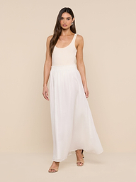 A woman with long, straight brown hair and a neutral expression stands against a white background, wearing a sleeveless beige top paired with the Evangelina Maxi Skirt—a long, breathable white summer skirt—and sandals.