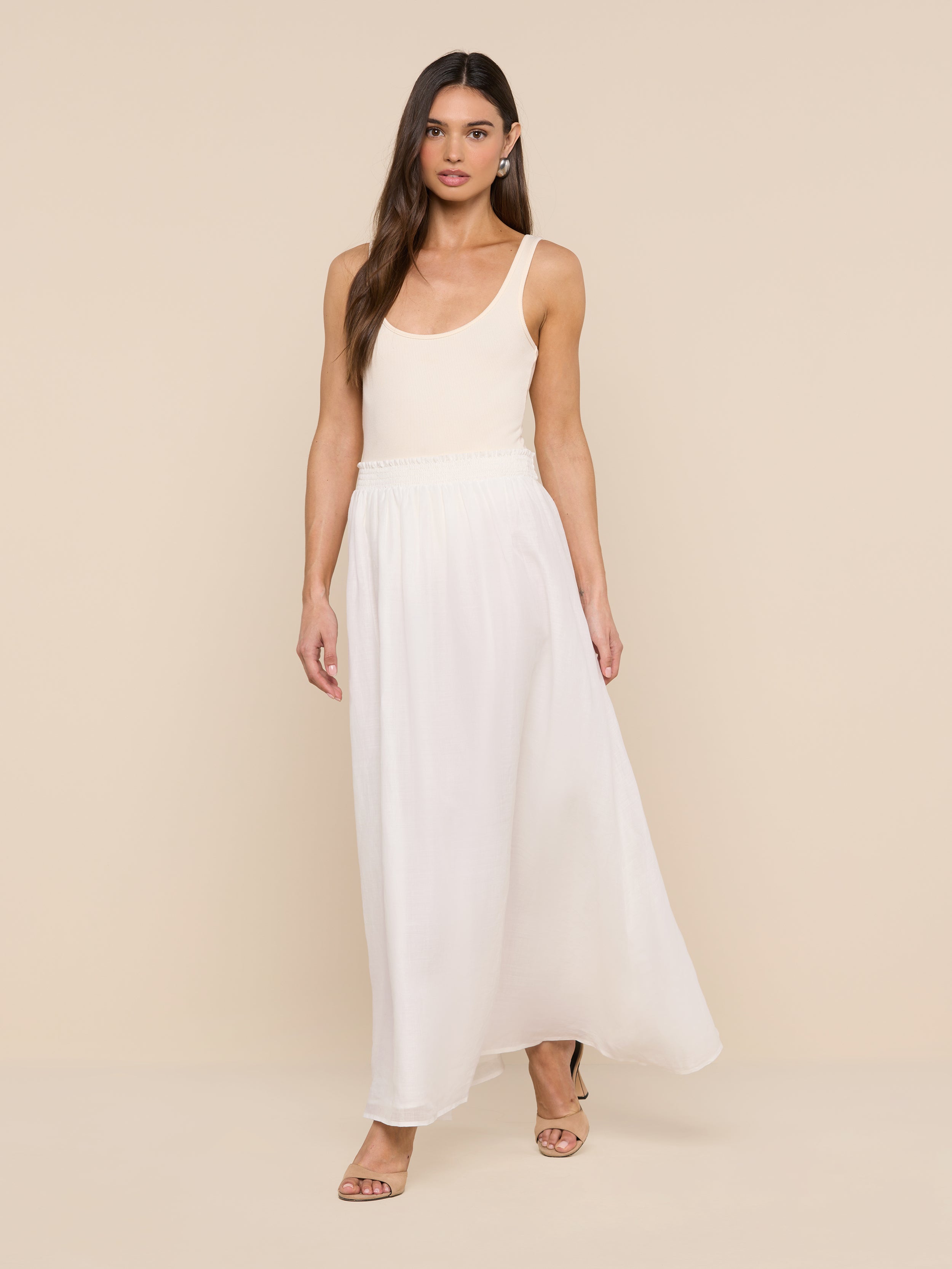 A woman with long, straight brown hair and a neutral expression stands against a white background, wearing a sleeveless beige top paired with the Evangelina Maxi Skirt—a long, breathable white summer skirt—and sandals.