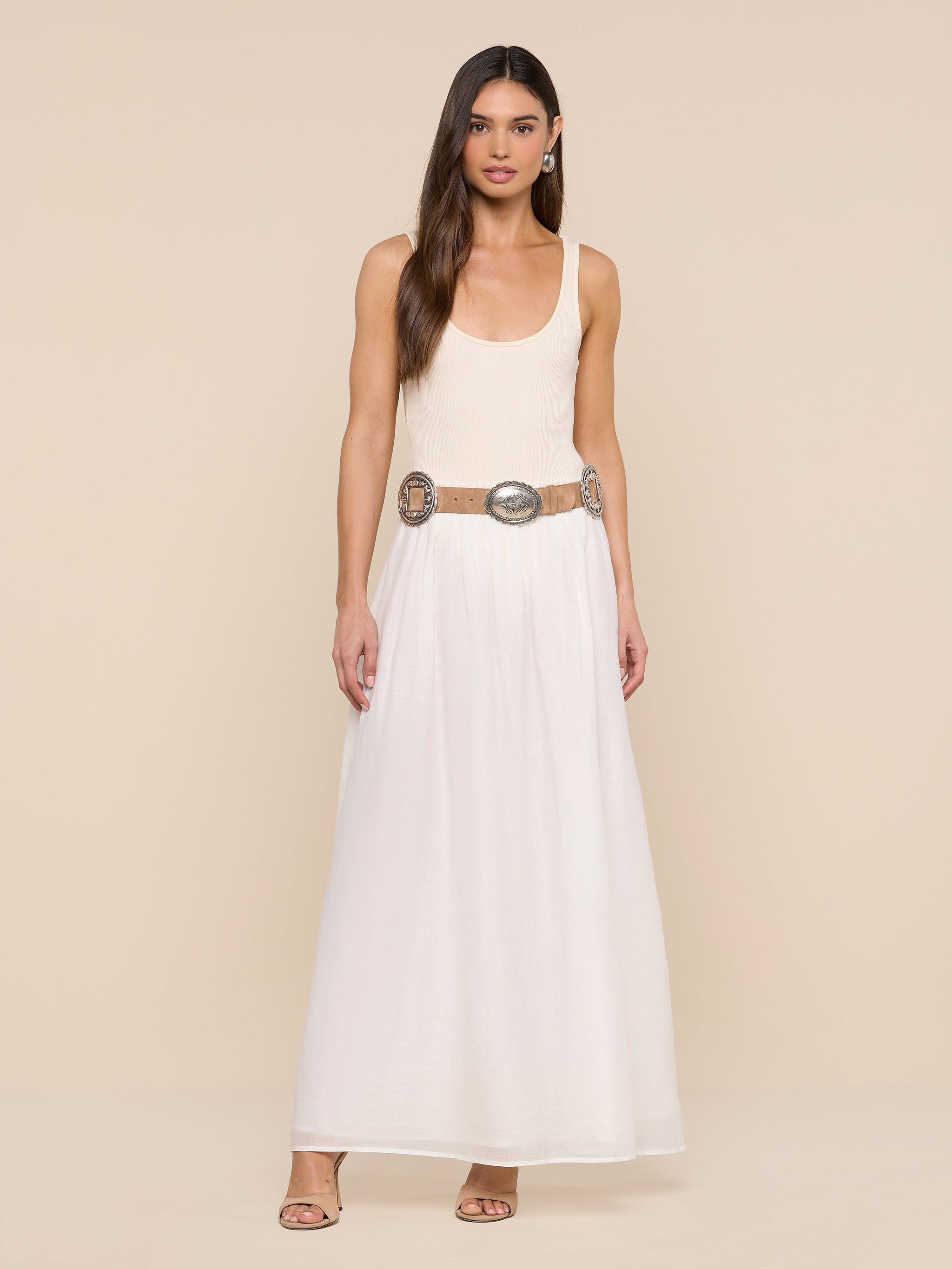 A woman stands against a white background wearing a sleeveless beige top, the Evangelina Maxi Skirt—a long, breathable white summer skirt—a wide decorative belt with large round details, hoop earrings, and open-toe shoes.