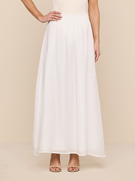 A person stands on a white background, shown from the mid-torso down, wearing the Evangelina Maxi Skirt—a long, flowing white summer skirt—paired with a sleeveless beige top. One arm is bent with their hand resting on their hip.