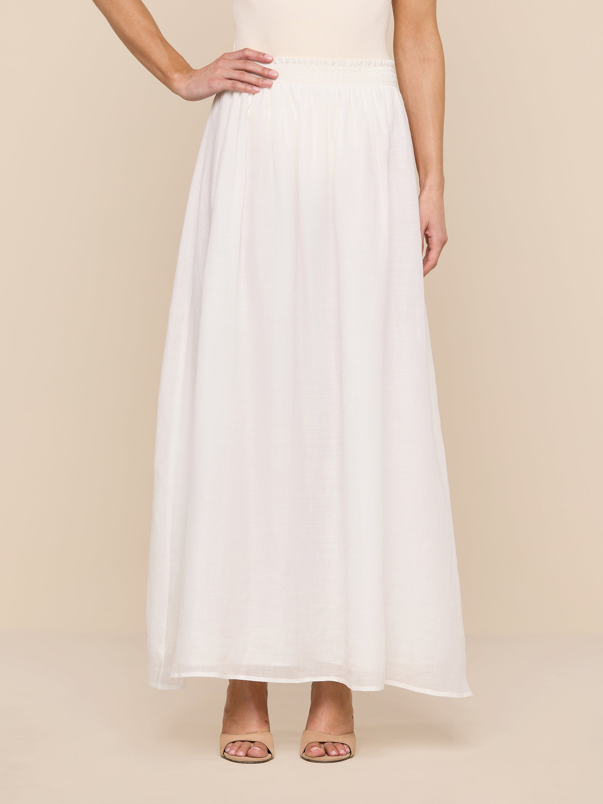 A person stands on a white background, shown from the mid-torso down, wearing the Evangelina Maxi Skirt—a long, flowing white summer skirt—paired with a sleeveless beige top. One arm is bent with their hand resting on their hip.